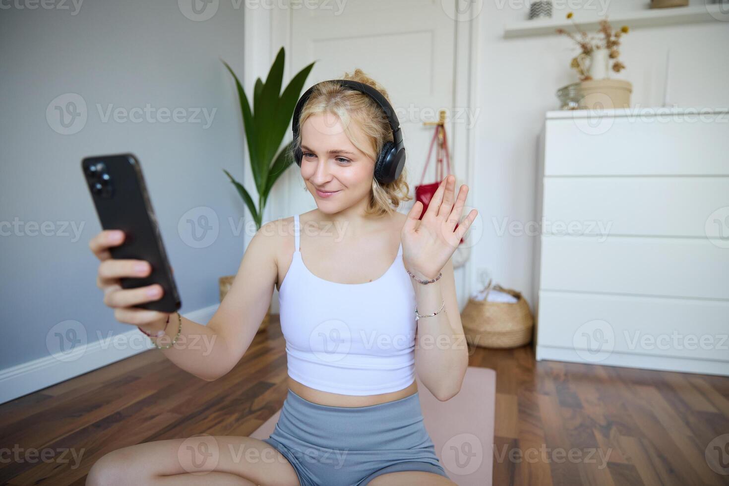 Portrait of young sporty woman in headphones, records on her smartphone, live streaming and saying hello to followers while doing workout training at home photo