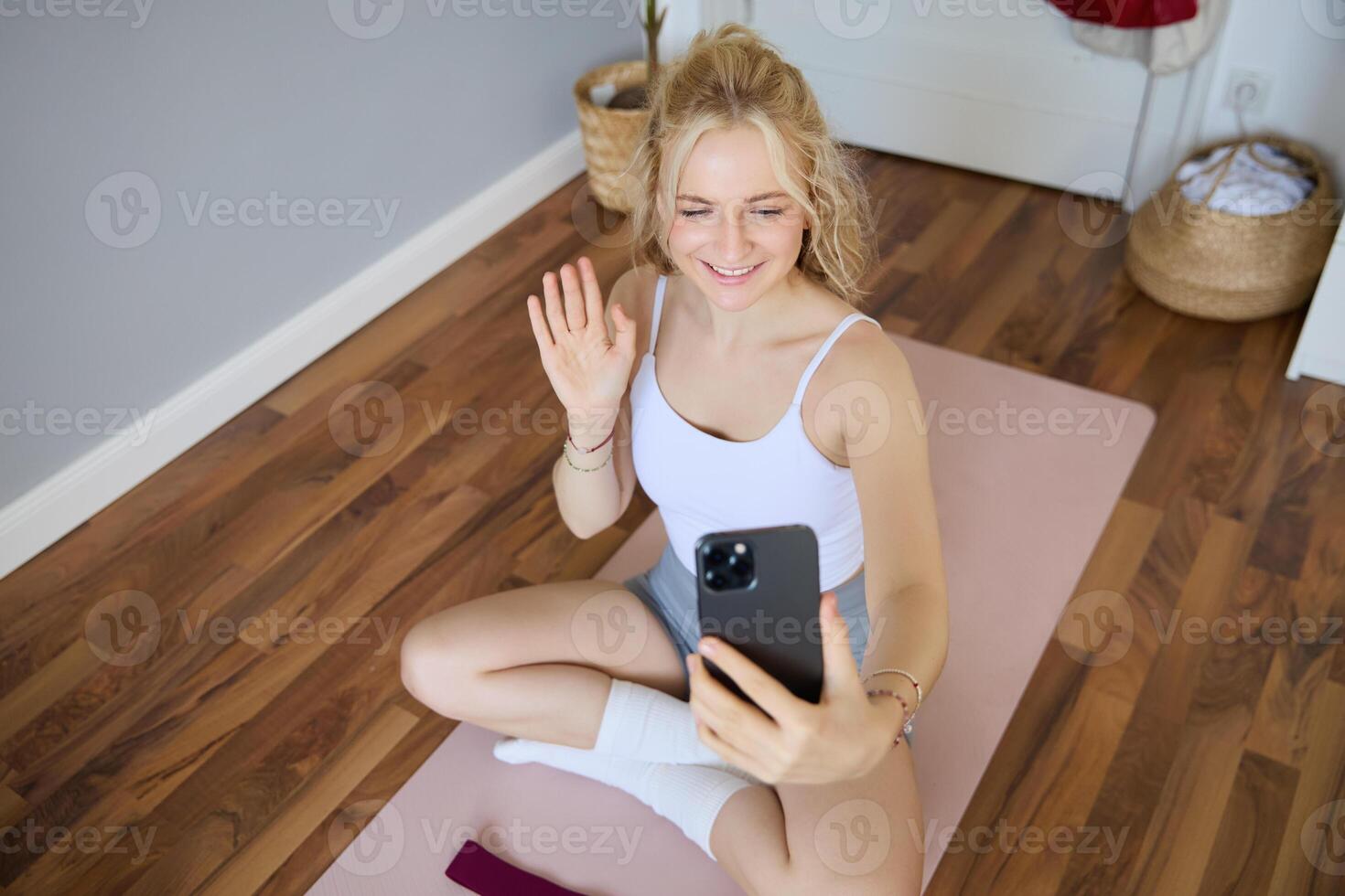retrato de adaptar, deportivo joven aptitud instructor, mujer da en línea rutina de ejercicio sesiones a clientela, olas mano a teléfono inteligente, se sienta en yoga estera y muestra ejercicios foto