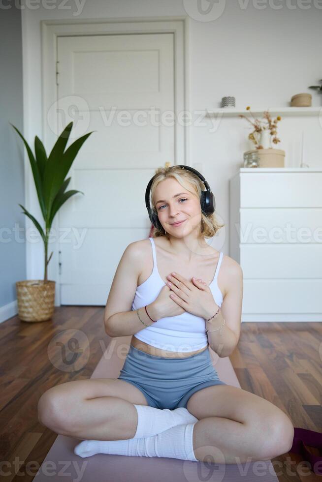 retrato de joven rubio mujer en auriculares, sensación calma y relajado después meditación, yoga capacitación, sentado en yoga estera en su habitación foto