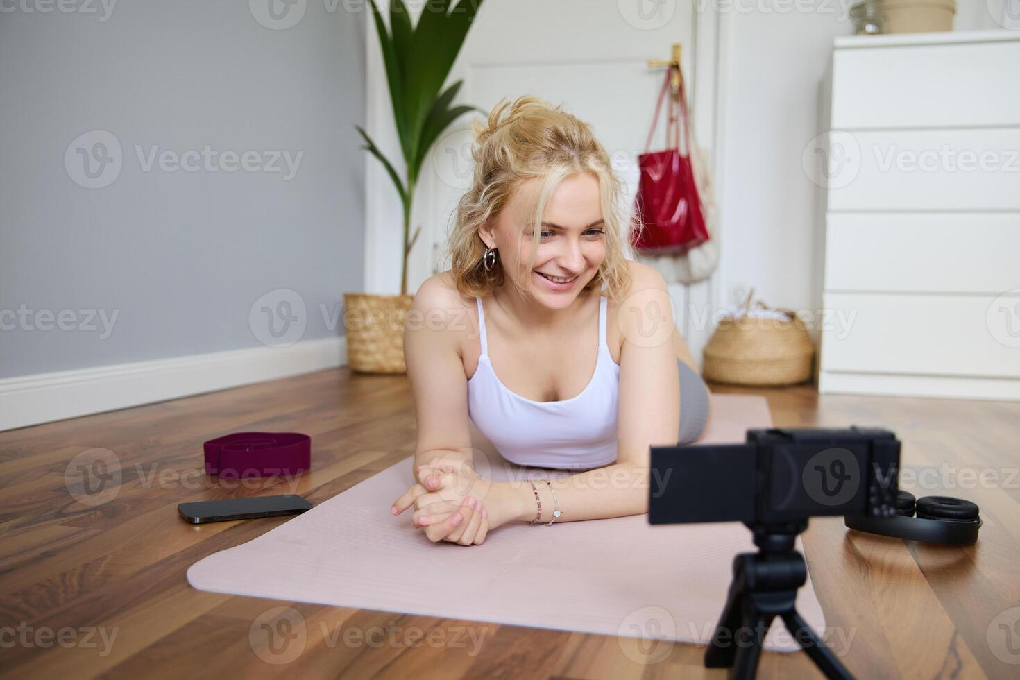 Portrait of young fitness instructor, vlogger creating content at home, doing workout and record exercises on digital camera, using rubber yoga mat photo