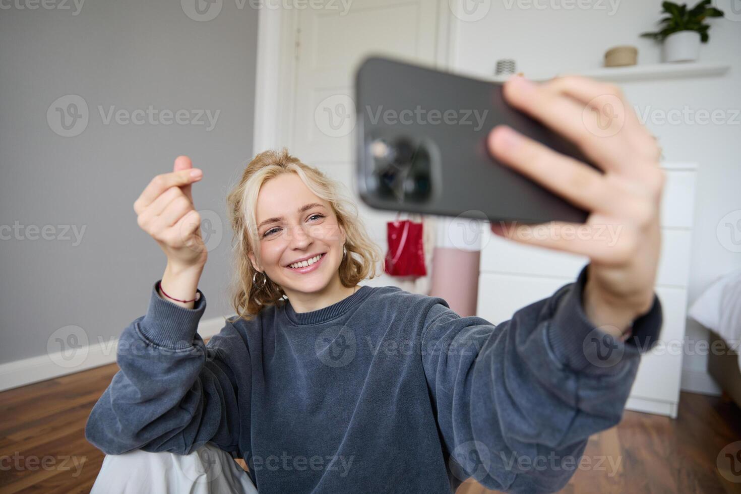 Image of young woman, vlogger taking selfie in her room, talking to her followers during online live stream, using smartphone app to chat with audience, smiling and looking happy photo