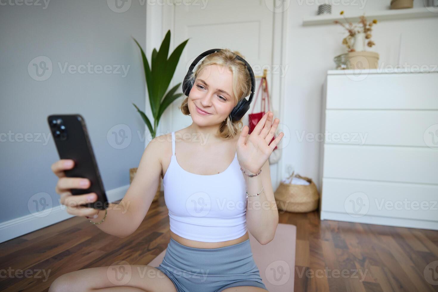 Portrait of young woman with smartphone and headphones, waving hand at mobile phone camera, live streaming during workout photo