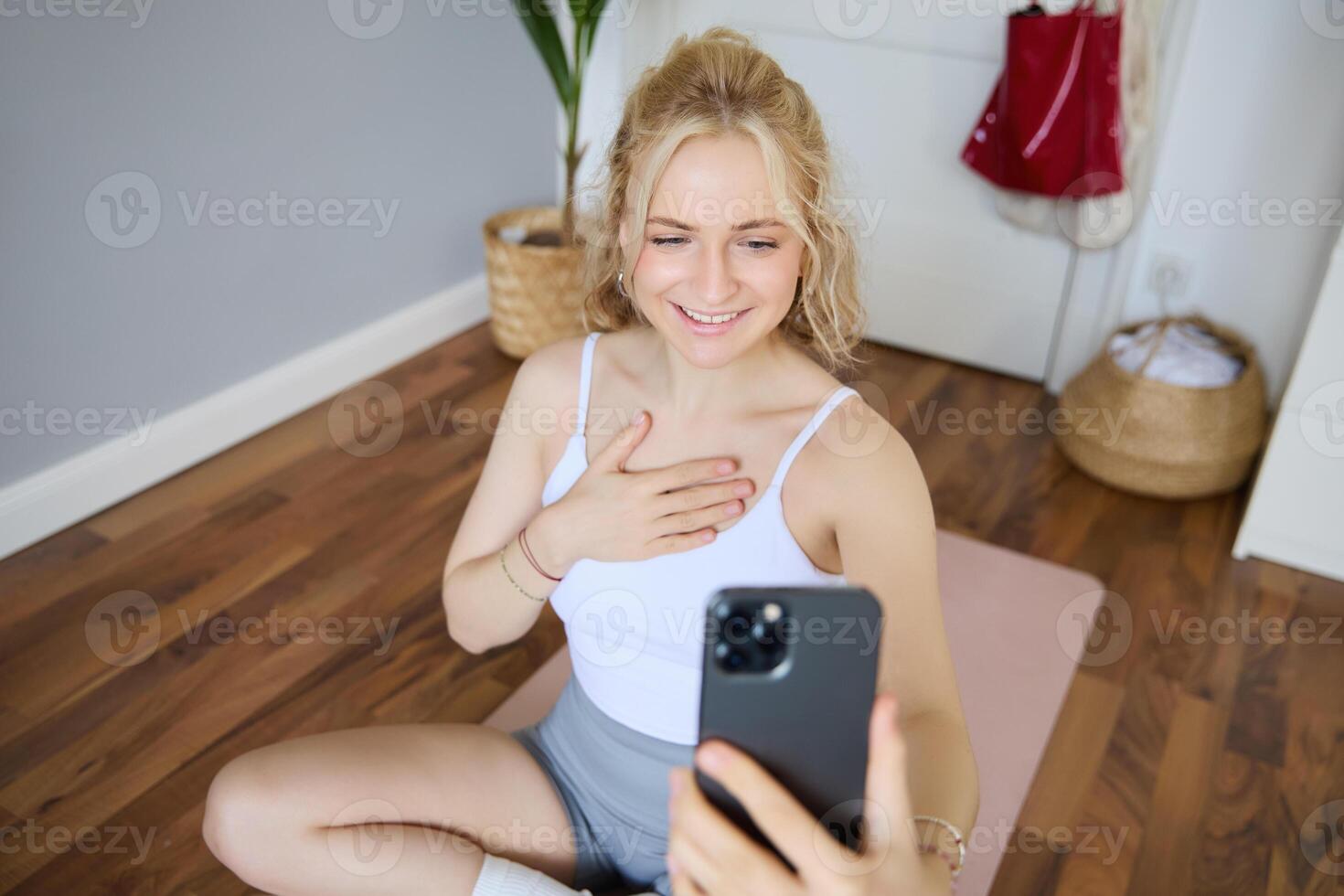 retrato de joven hermosa rubio mujer, aptitud instructor haciendo ejercicios a hogar en yoga estera, tomando selfies en teléfono inteligente, grabación de sí misma para social medios de comunicación cuenta acerca de rutina de ejercicio foto