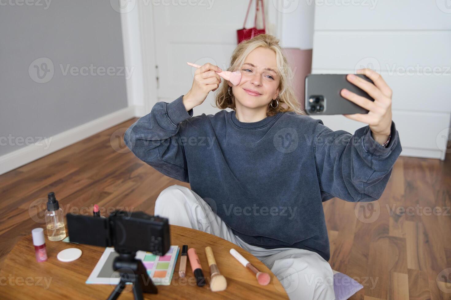 Portrait of beautiful young lifestyle blogger, woman vlogger recording on digital camera, live stream makeup tutorial on smartphone app, sitting in bedroom, using brush on her cheeks photo