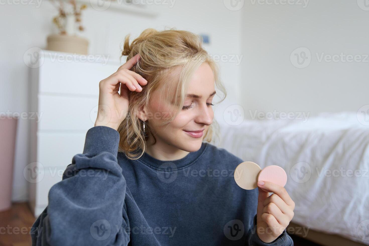 Portrait of young woman chatting on live stream about makeup, sits on floor in bedroom, showing beauty products to followers photo