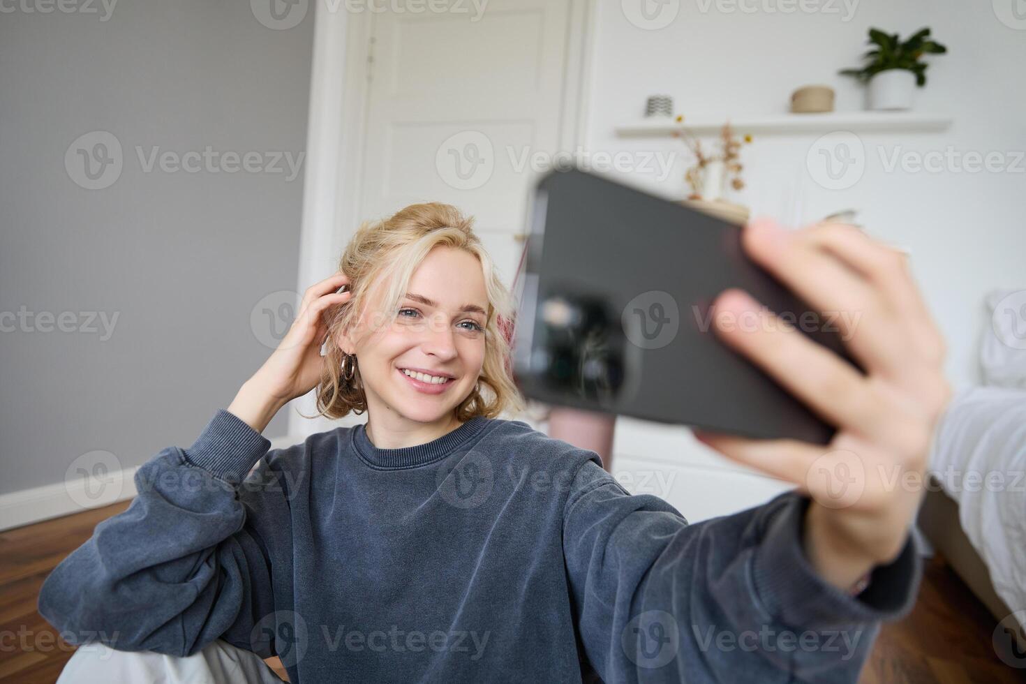 Portrait of young woman, social media influencer, taking selfies in her room, sitting on floor, holding smartphone and posing for a photo