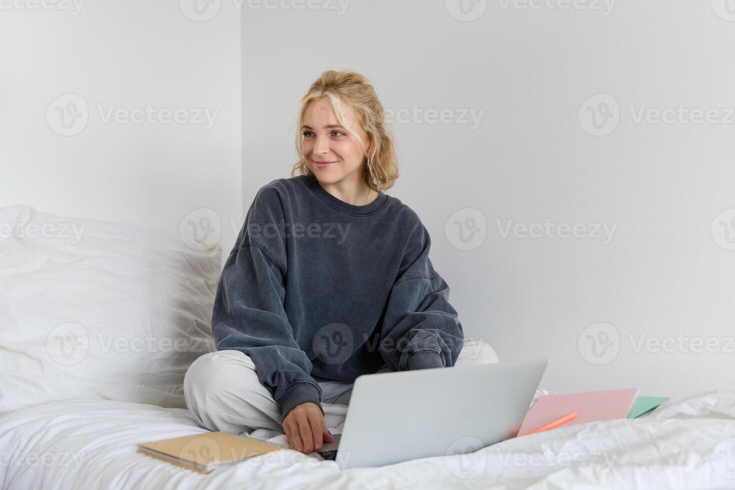 Portrait of happy blond woman, freelancer working from home, sitting on bed with laptop and notebooks. Student doing homework in bedroom, connects to online class via chat photo