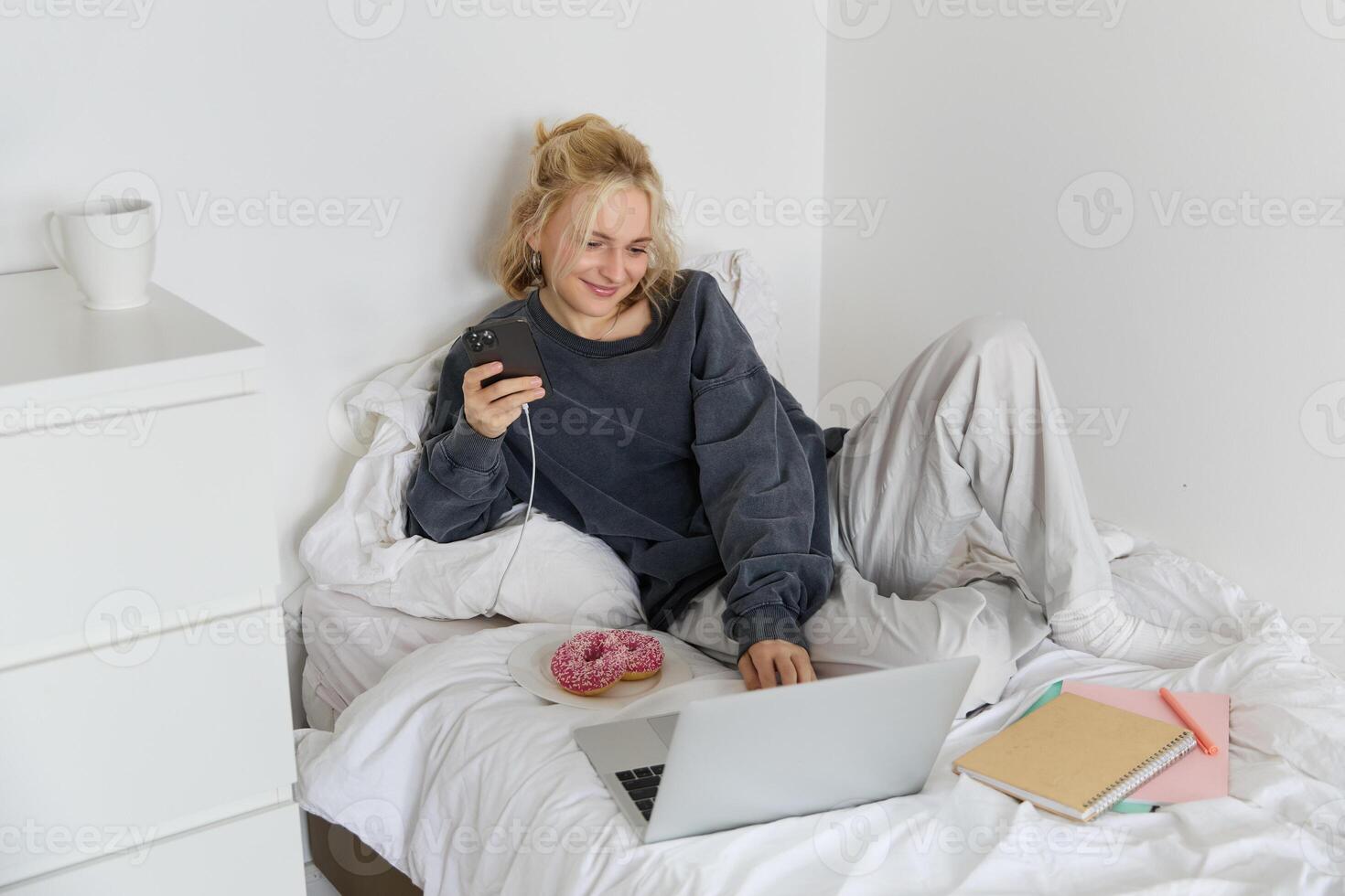 Lifestyle and weekend concept. Young smiling woman, relaxing at home, lying in bed with cup of tea and doughnut, using laptop and smartphone in bedroom photo