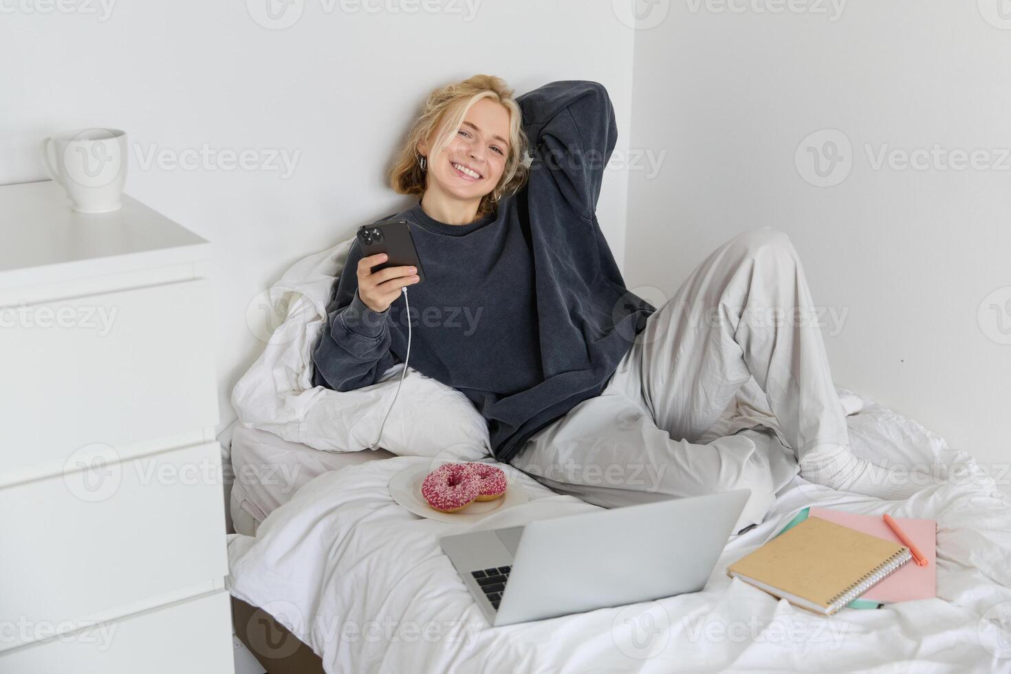 Portrait of young beautiful blond woman, feeling lazy at home, lying in bed with cup of tea, doughnut and laptop, studying remotely or watching movie online photo