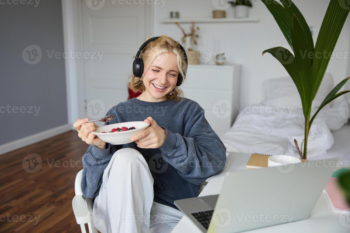 Portrait of smiling young woman, watching tv show in headphones, eating breakfast and looking at laptop screen photo