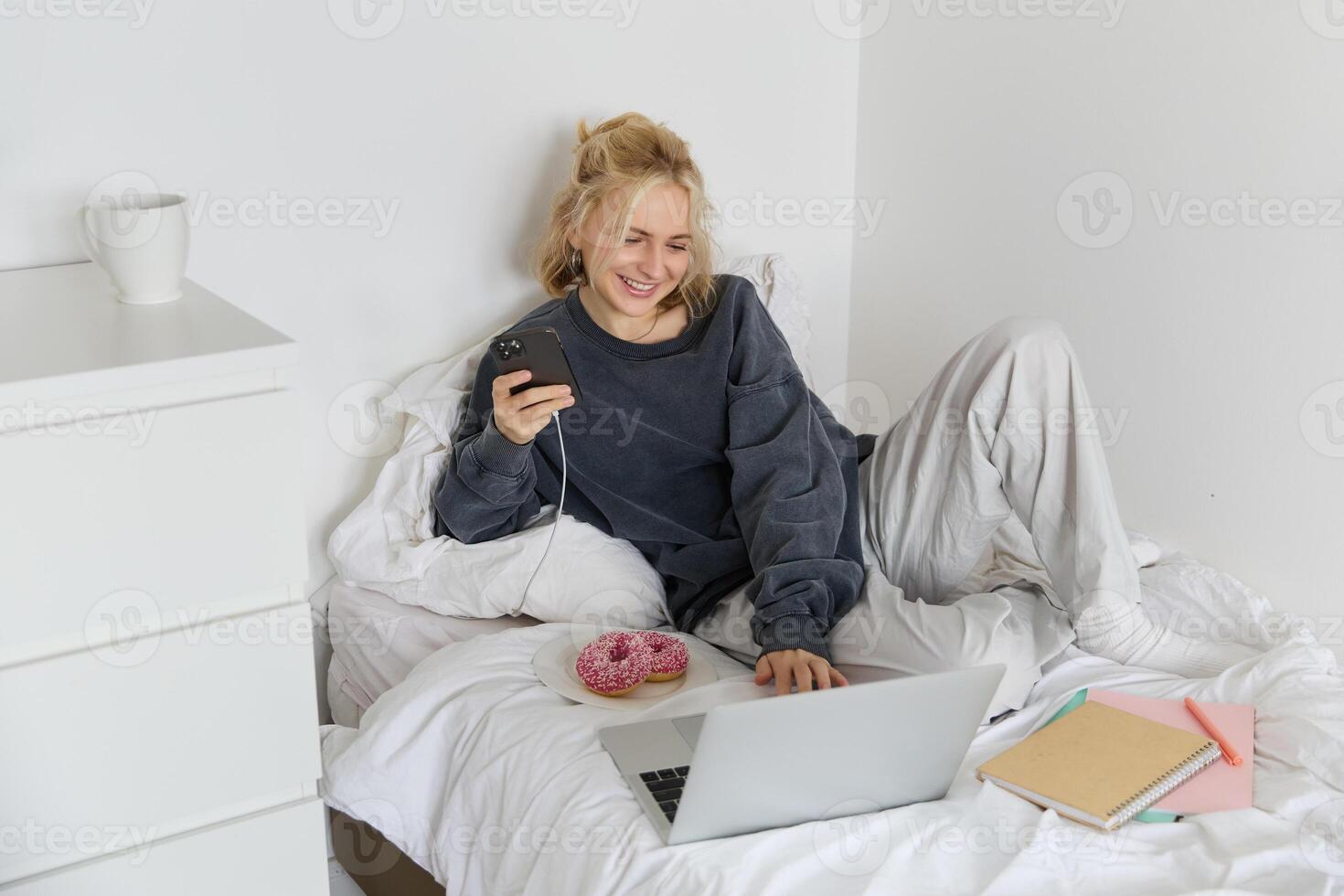 Lifestyle and people concept. Young beautiful woman, staying at home, lying in bed with laptop and smartphone, eating doughnut, enjoying free time, spending weekend at home, watching movie online photo