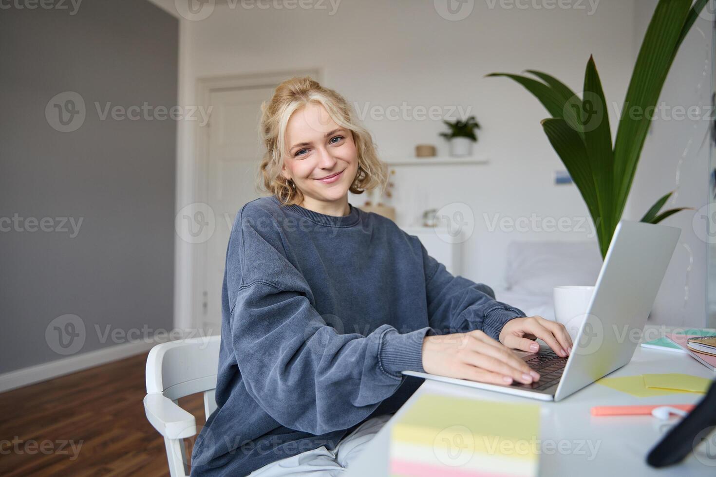 retrato de joven sonriente hermosa mujer, estudiante estudiando remotamente en su habitación, aprendizaje electrónico, utilizando computadora portátil, sentado en silla en dormitorio, mirando contento a cámara foto