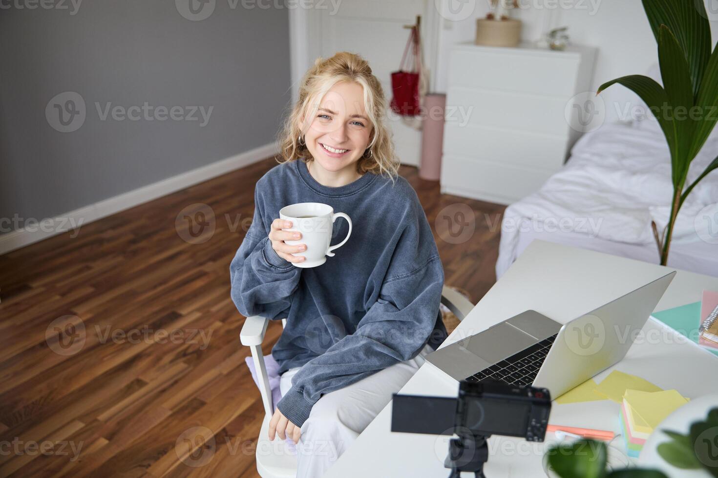 Portrait of young smiling woman, vlogger recording lifestyle in her room, sits in front of laptop and digital camera, drinks tea, creates content for social media account photo
