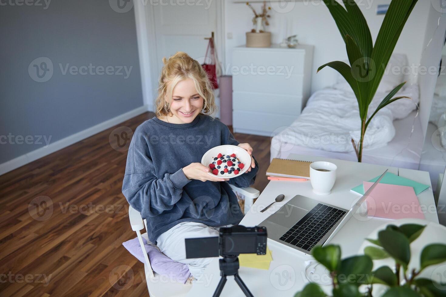Portrait of cute young girl vlogger, showing her breakfast on camera, recording vlog in her bedroom, holding dessert, eating photo