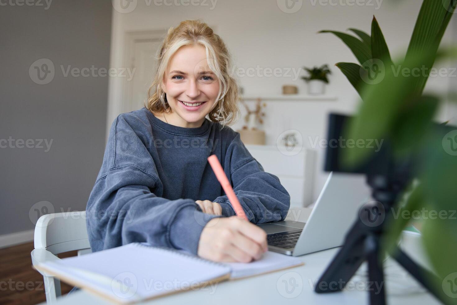 Cute smiling girl sits in a room, writes down notes, doing homework, records of herself on digital camera, creates content for vlog, lifestyle blogger doing daily routine episode photo