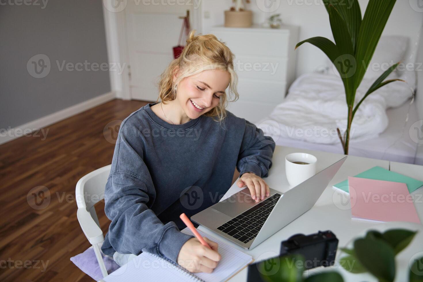 retrato de sonriente, hermosa rubio mujer, escritura abajo notas, haciendo tarea, estudiando desde hogar, haciendo distancia aprendiendo, en línea curso, trabajando remotamente en su habitación foto