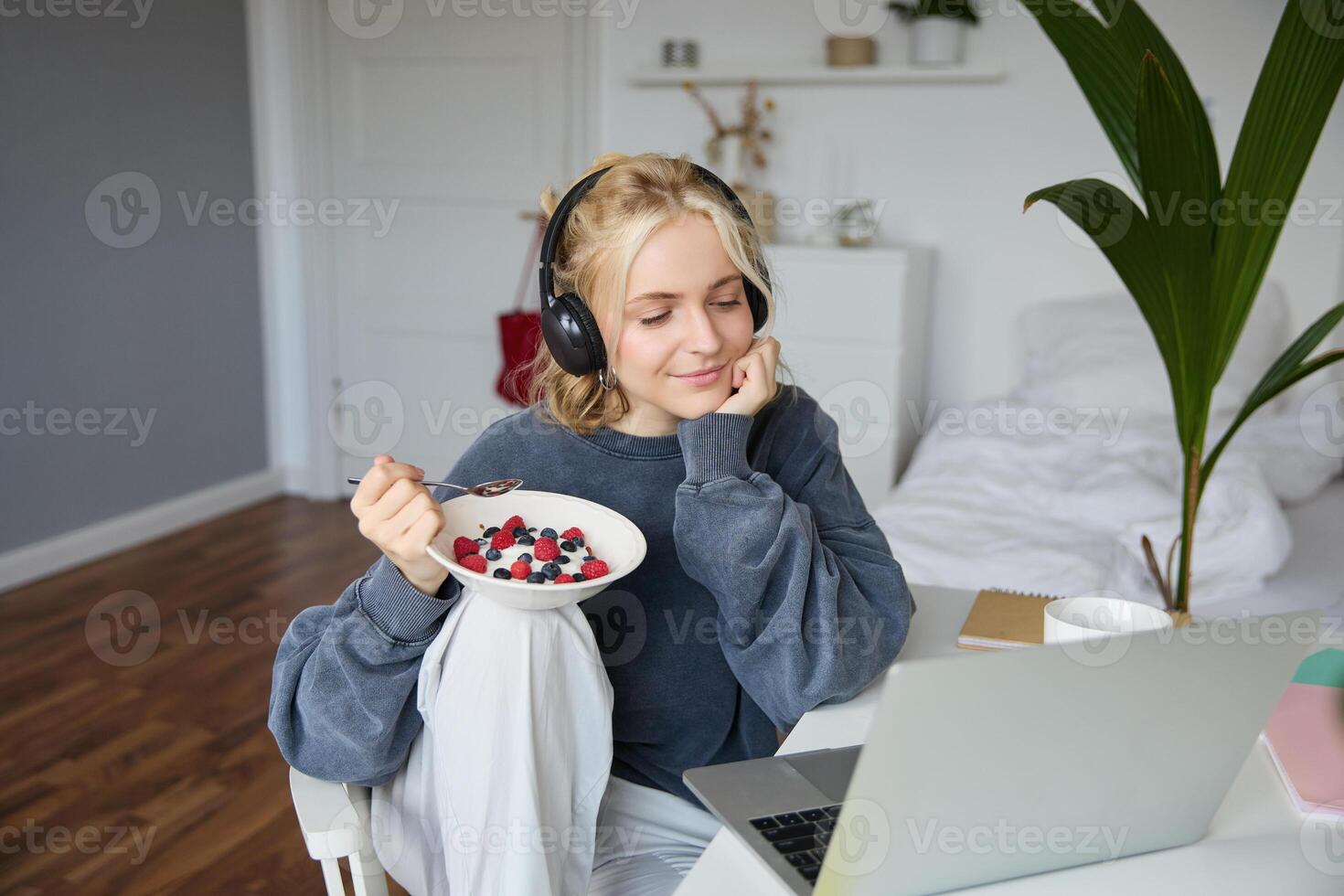 Image of happy woman sitting in a room, watching interesting tv show or movie on laptop, using screaming service, wearing headphones, eating dessert and drinking tea photo
