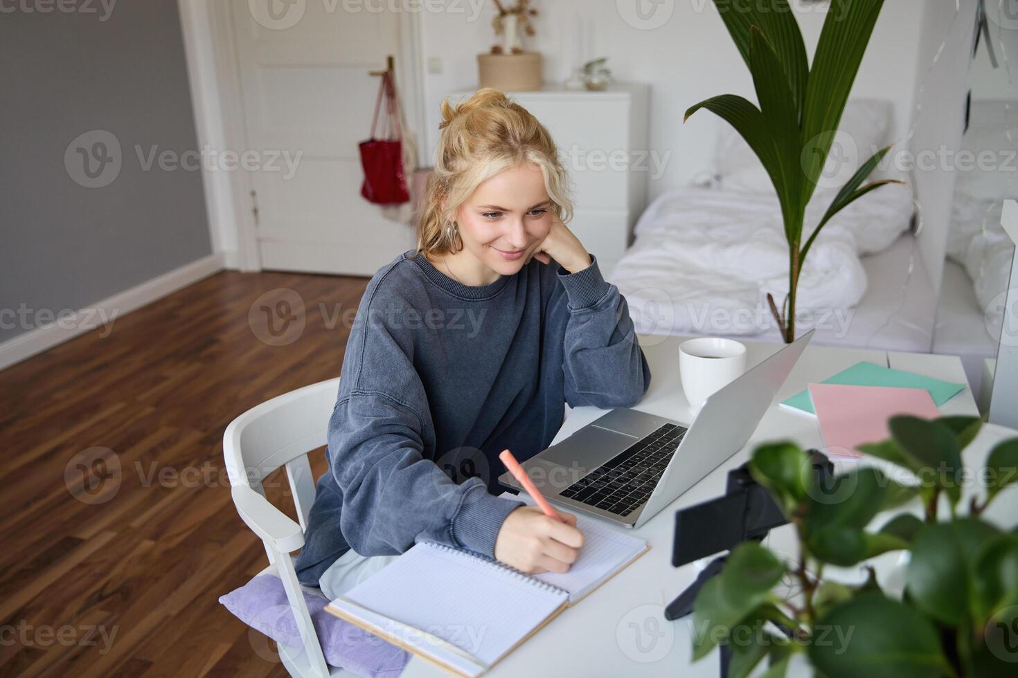 retrato de sonriente, hermosa rubio mujer, escritura abajo notas, haciendo tarea, estudiando desde hogar, haciendo distancia aprendiendo, en línea curso, trabajando remotamente en su habitación foto