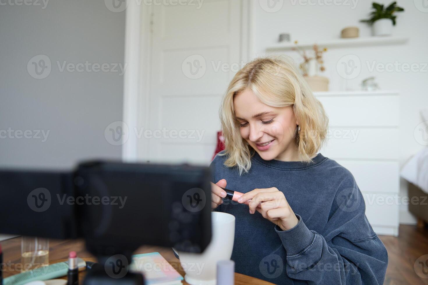 Close up portrait of smiling young woman with camera in her room, showing mascara, reviewing beauty products for social media account. Beauty blogger creates vlog about makeup photo