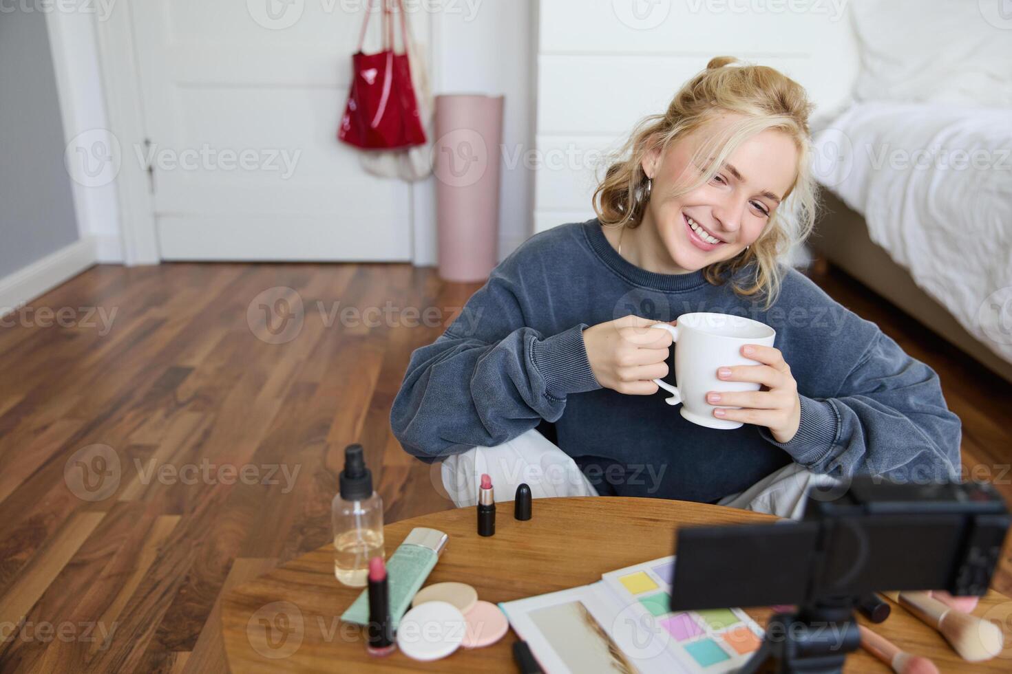 Young woman smiling, recording lifestyle vlog on her digital camera, holding cup of tea while talking, has makeup on coffee table, doing makeup tutorial for social media from her room photo