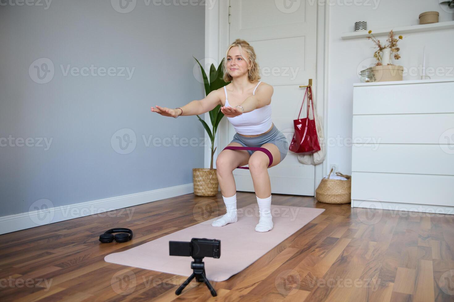 retrato de joven mujer, aptitud instructor haciendo un para deporte vlog, haciendo sentadillas en cámara, utilizando resistencia banda, trabajando fuera a hogar foto