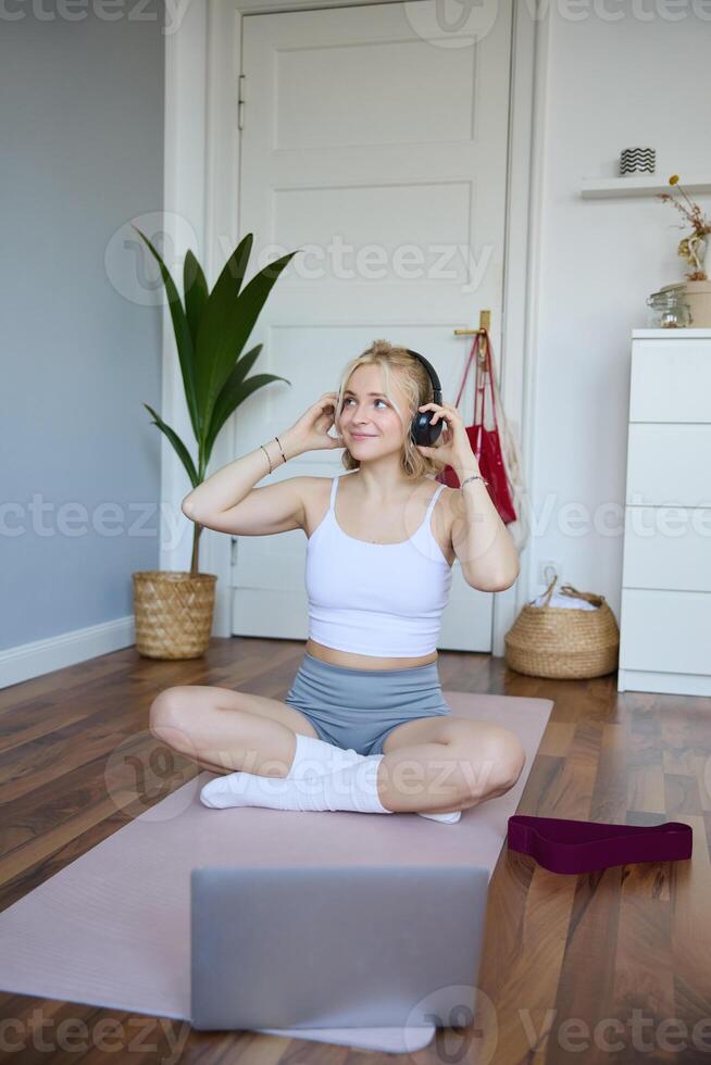 vertical Disparo de sonriente joven mujer utilizando tutoriales para rutina de ejercicio a hogar, sentado con ordenador portátil en inalámbrico auriculares en yoga estera, sigue aptitud instructor en social medios de comunicación, se repite ejercicios foto