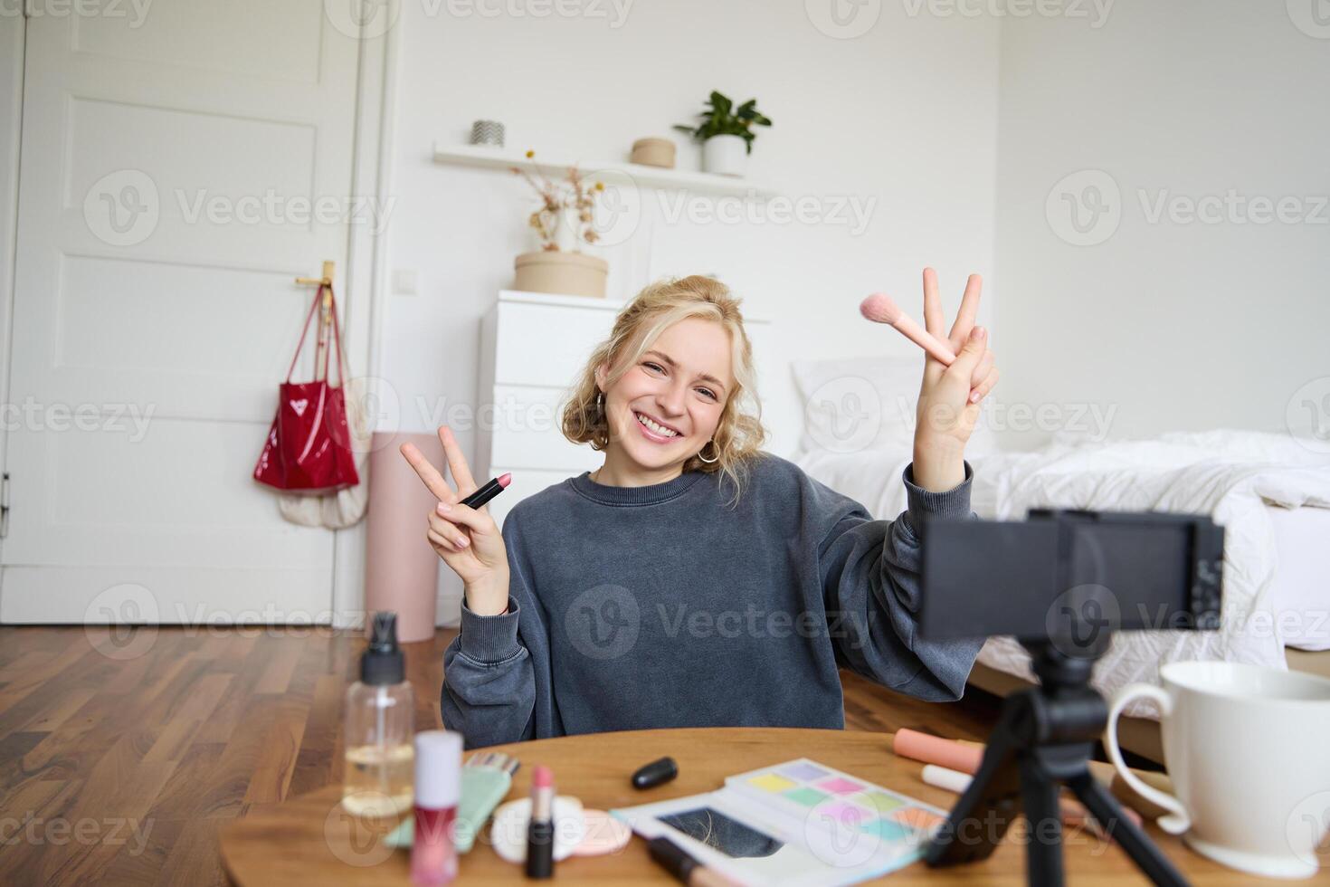 Portrait of young blond woman, teenage girl records for her social media account, shows makeup on camera, recommends lipstick to online followers, creates content in her room photo