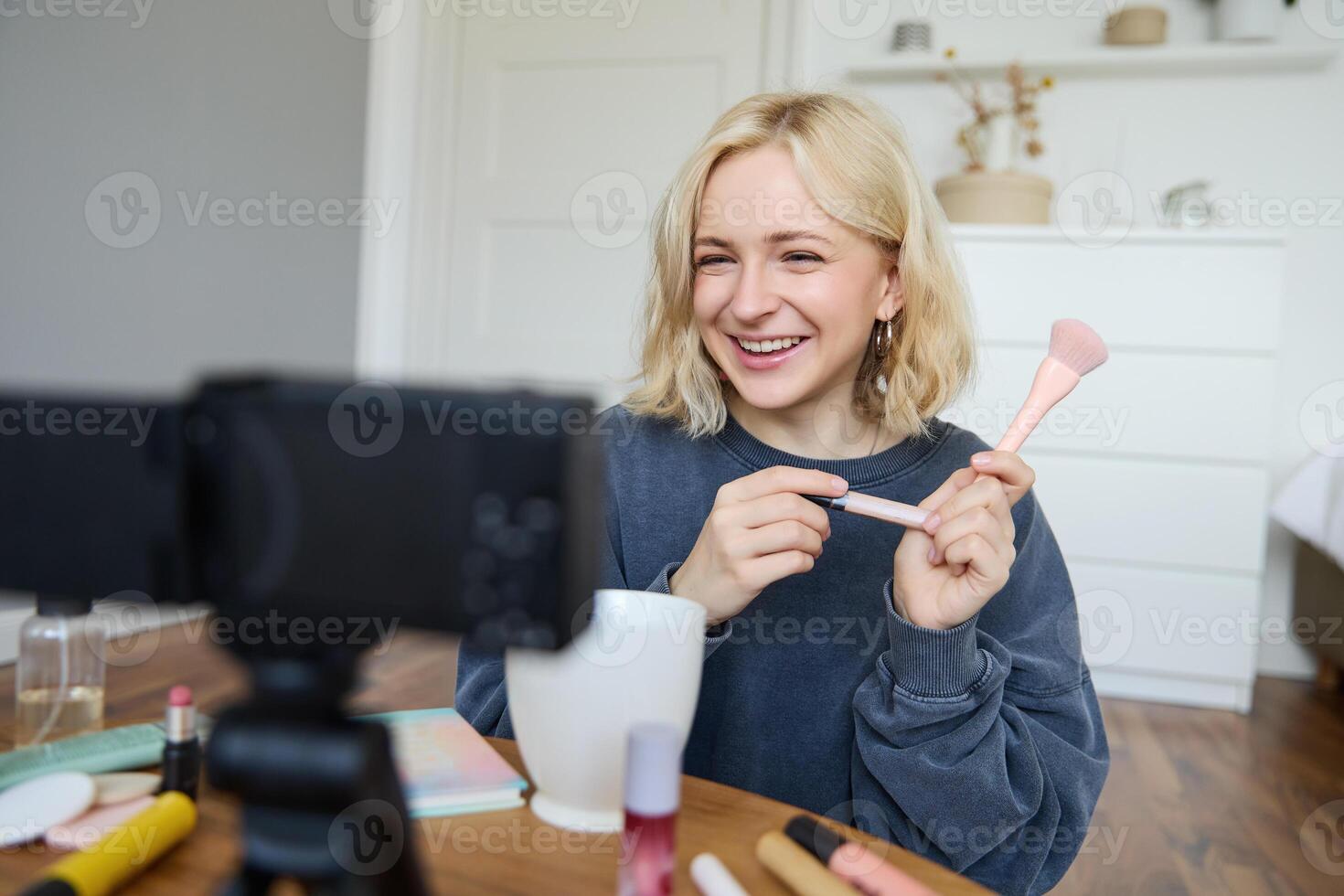 Close up portrait of happy young beauty blogger, records lifestyle vlog in her room, using camera with stabiliser, shows makeup brush and cosmetics photo