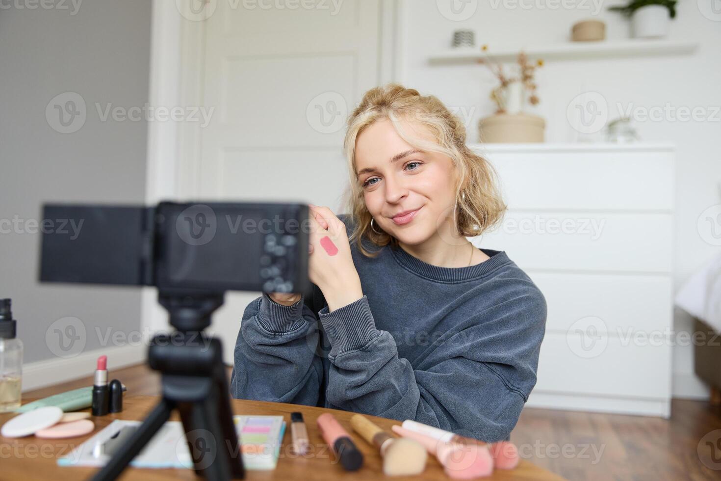 imagen de joven elegante mujer, blogger grabación un belleza estilo de vida de su cosecha mejor lápiz labial, demostración labio bálsamo muestras en su piel, sentado en frente de digital cámara en vacío habitación foto