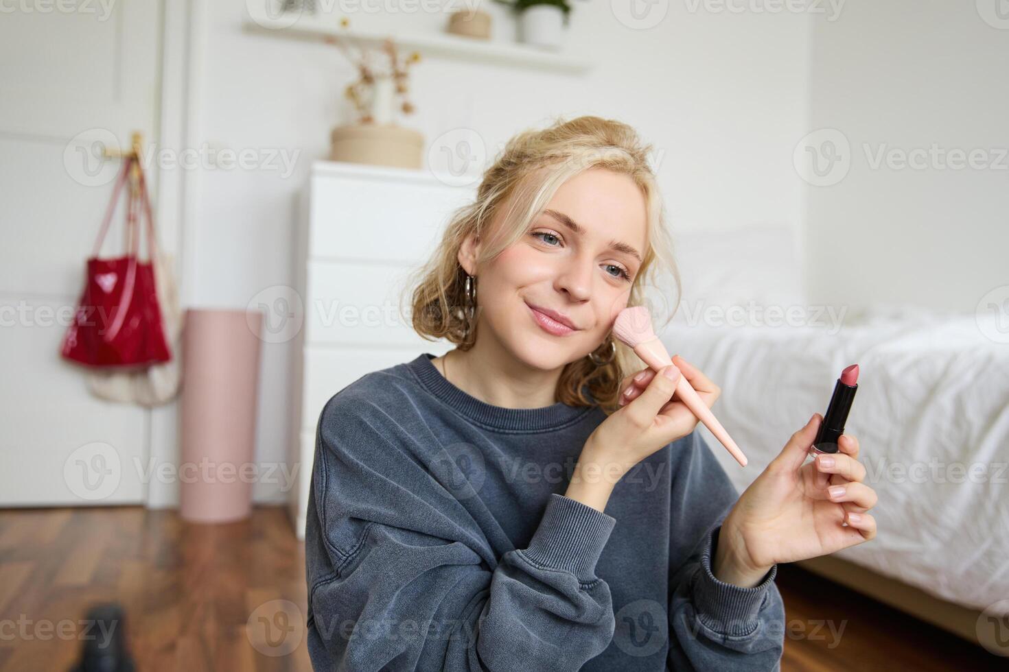 Woman beauty blogger, records of herself sitting in a room and rating makeup products, puts on make up, holds lipstick and cosmetic brush in hand, using professional camera for content creation photo