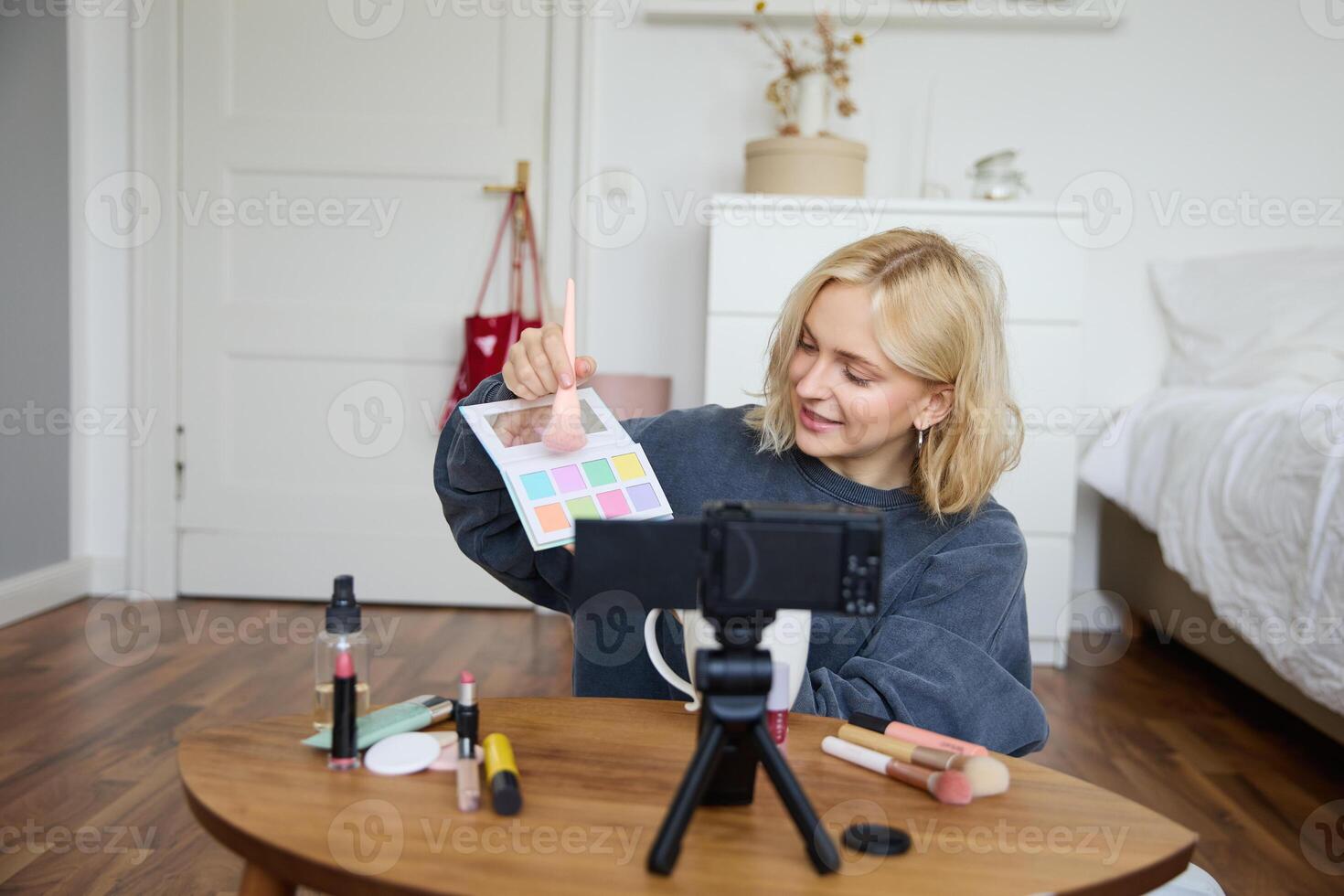 retrato de sonriente rubio mujer, Adolescente niña registros un vlog en cámara, vlogger muestra sombras de ojos, tutorial cómo a poner maquillaje en, se sienta en su habitación en piso cerca café mesa foto