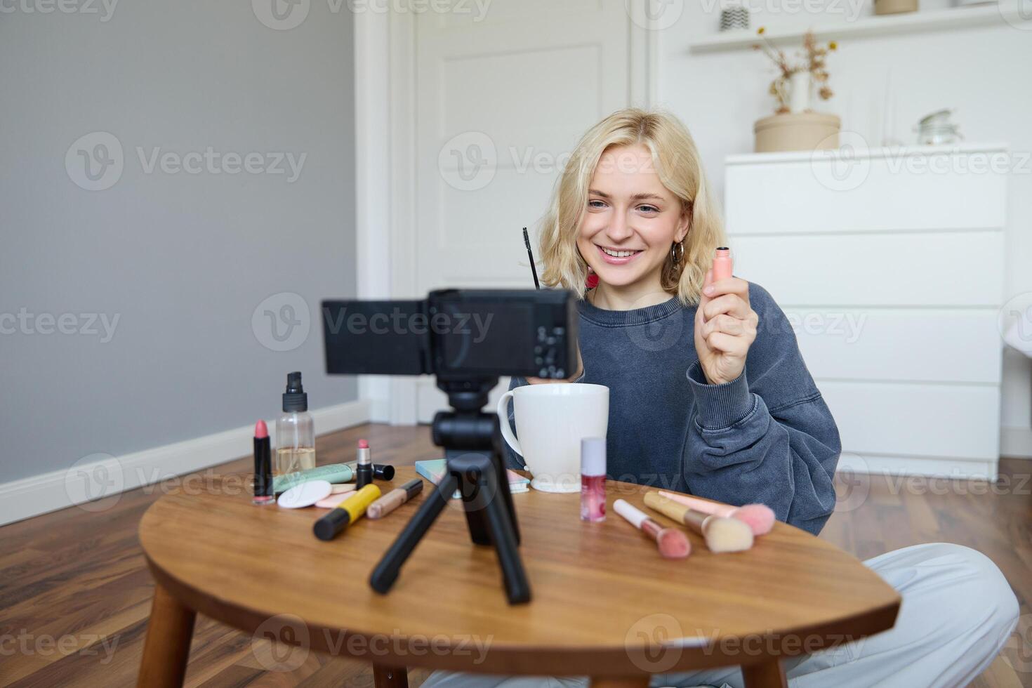 retrato de hermoso, sonriente rubio mujer, niña grabación de su maquillaje tutorial para social medios de comunicación, vlogger sentado en piso en su habitación, utilizando estabilizador a crear contenido, revisando máscara foto