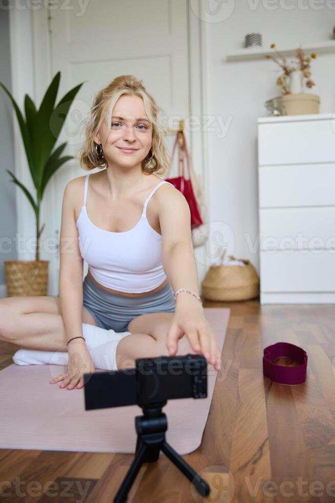 Vertical shot of woman sitting on yoga mat, recording on digital camera, vlogging about workout at home and fitness lifestyle, showing how to stay fit and healthy indoors photo