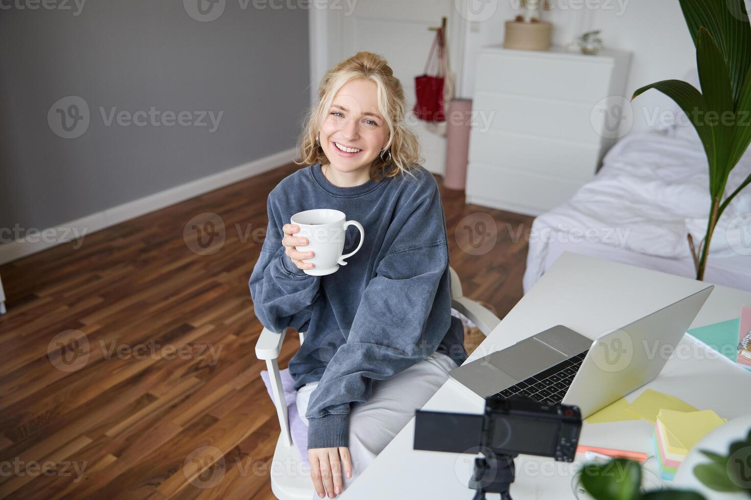 Portrait of young smiling woman, vlogger recording lifestyle in her room, sits in front of laptop and digital camera, drinks tea, creates content for social media account photo
