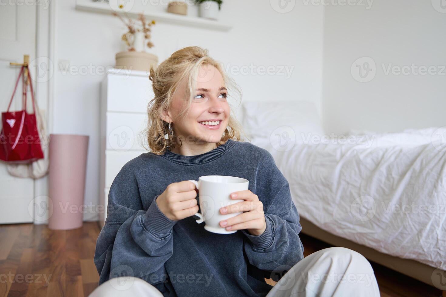 Lifestyle portrait of young woman sitting on bedroom floor with cup of tea, drinking from big white mug and looking aside, smiling happily photo