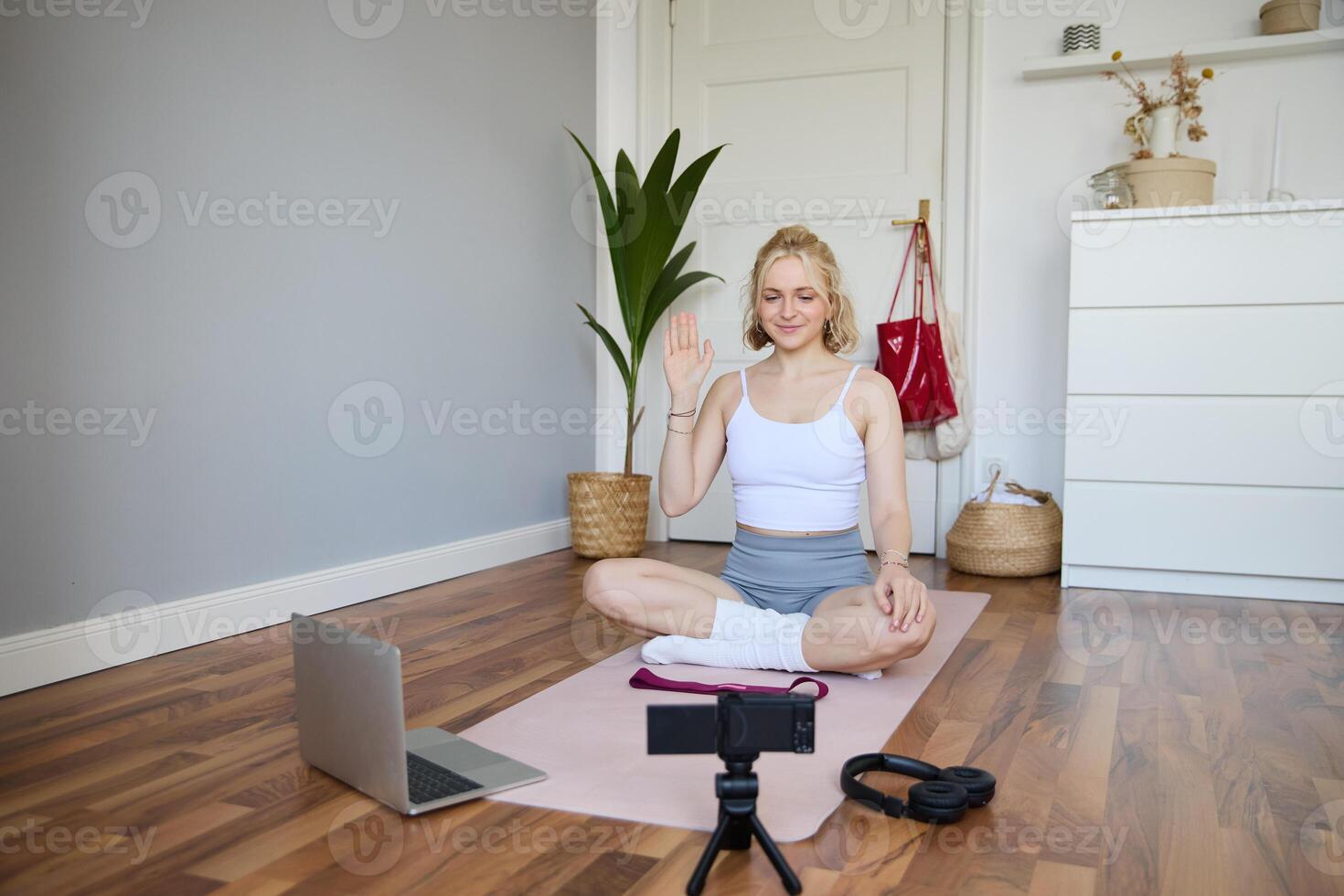 Portrait of young female athlete, fitness trainer recording vlog, training session on digital camera, sitting in a room on rubber yoga mat, showing exercises photo