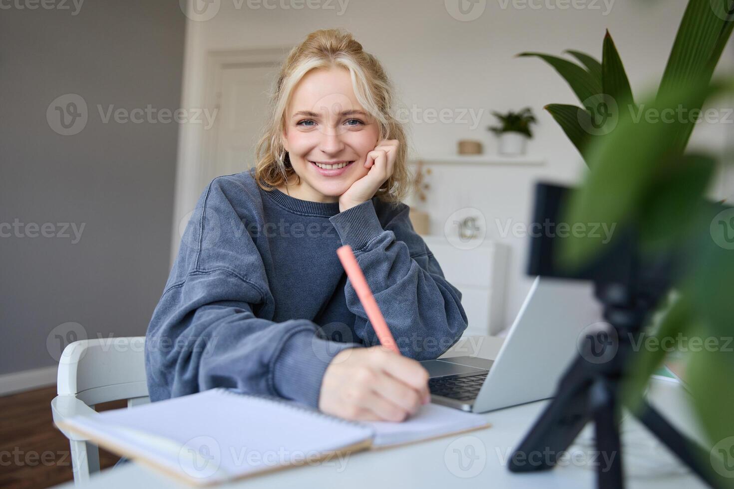 Cute smiling girl sits in a room, writes down notes, doing homework, records of herself on digital camera, creates content for vlog, lifestyle blogger doing daily routine episode photo
