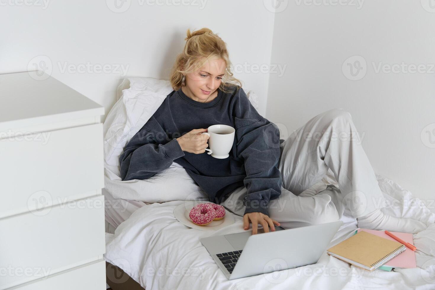 Portrait of young beautiful blond woman, feeling lazy at home, lying in bed with cup of tea, doughnut and laptop, studying remotely or watching movie online photo