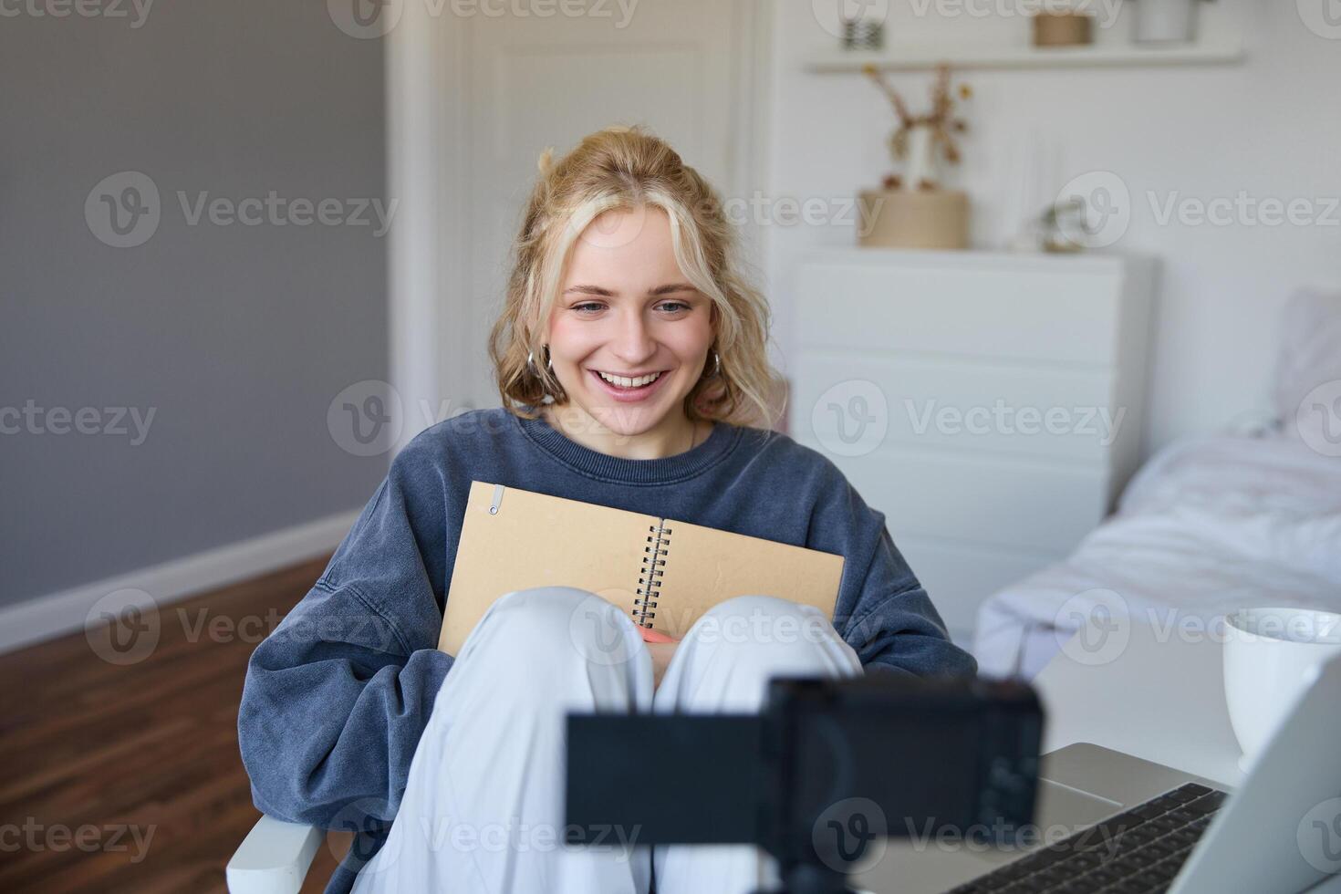 Portrait of blond smiling woman, records on digital camera how she writes in notebook, talks to followers, doing lifestyle blog content in her room photo