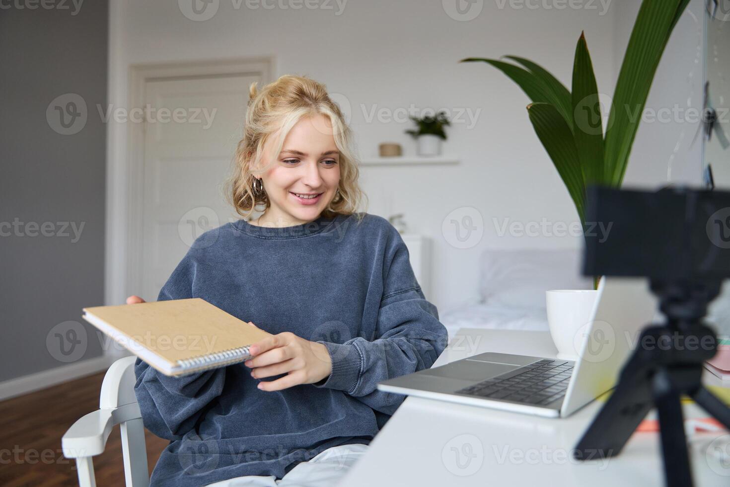 Young smiling blond woman, sits near laptop, uses digital camera to record blog, creates lifestyle content for social media, records tutorial, shows notebook, reads her notes photo