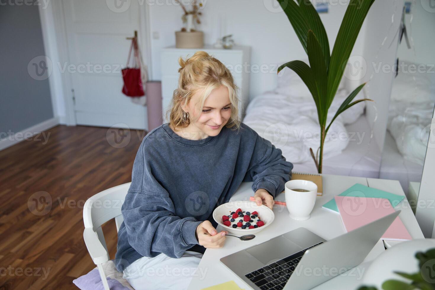 Portrait of smiling blond young woman, eating in front of laptop, watching s online while having breakfast, enjoying dessert, sitting in bedroom photo