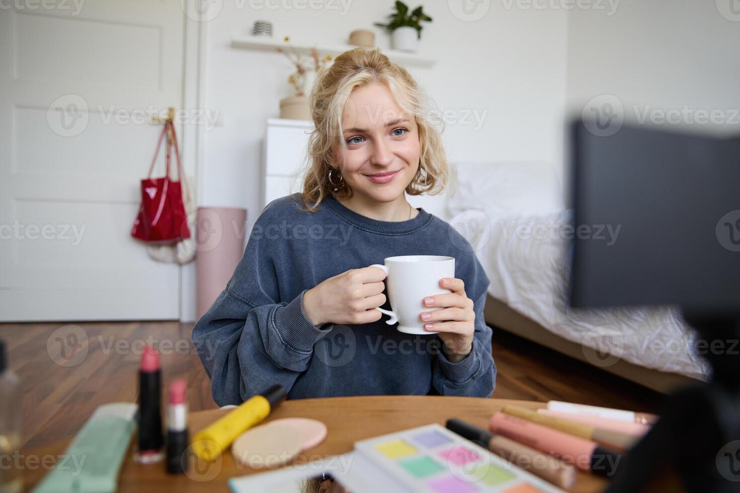 Portrait of cute blond woman, content maker, vlogger recording on digital camera in bedroom, drinking cup of tea and talking to audience photo