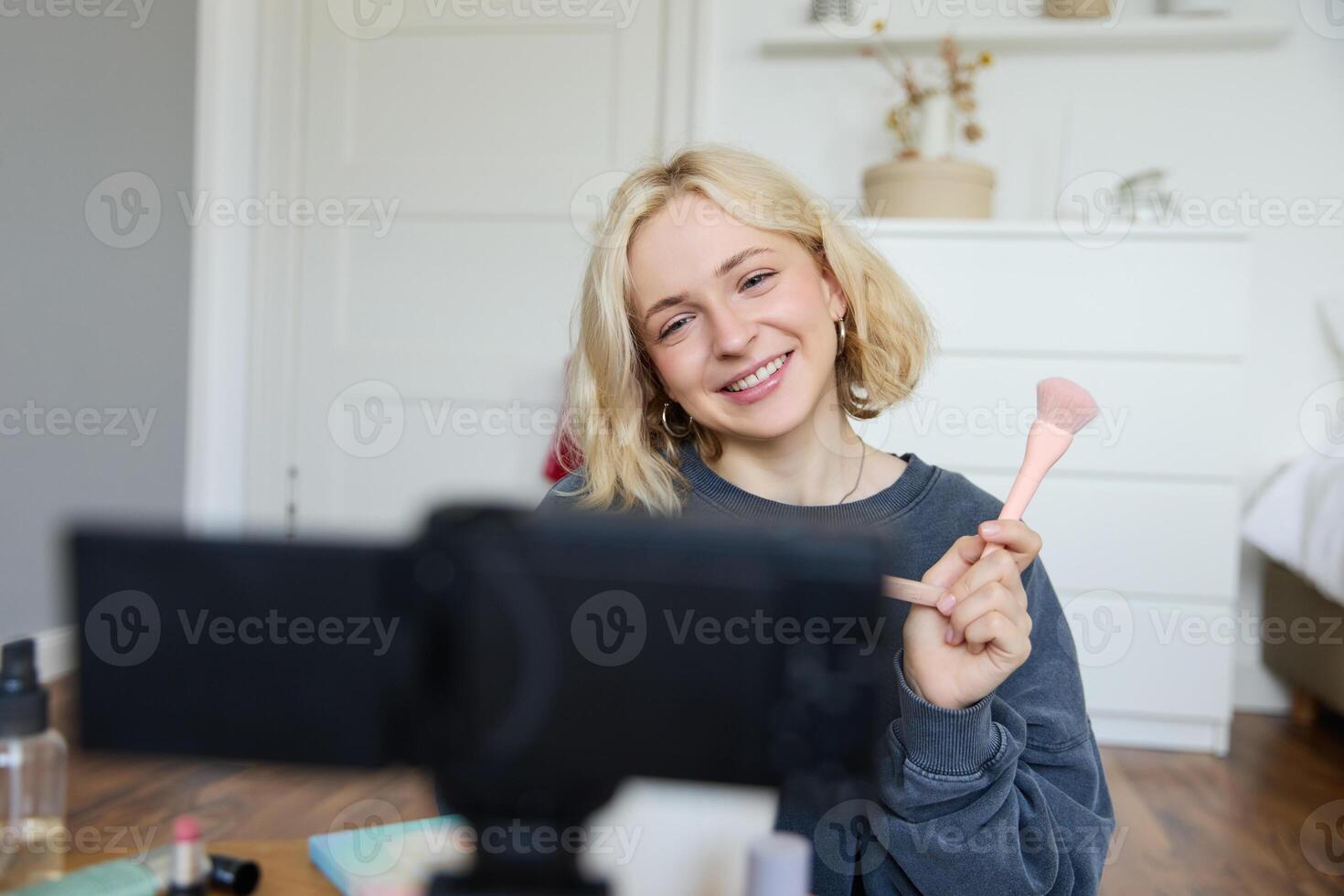 Close up portrait of happy young beauty blogger, records lifestyle vlog in her room, using camera with stabiliser, shows makeup brush and cosmetics photo