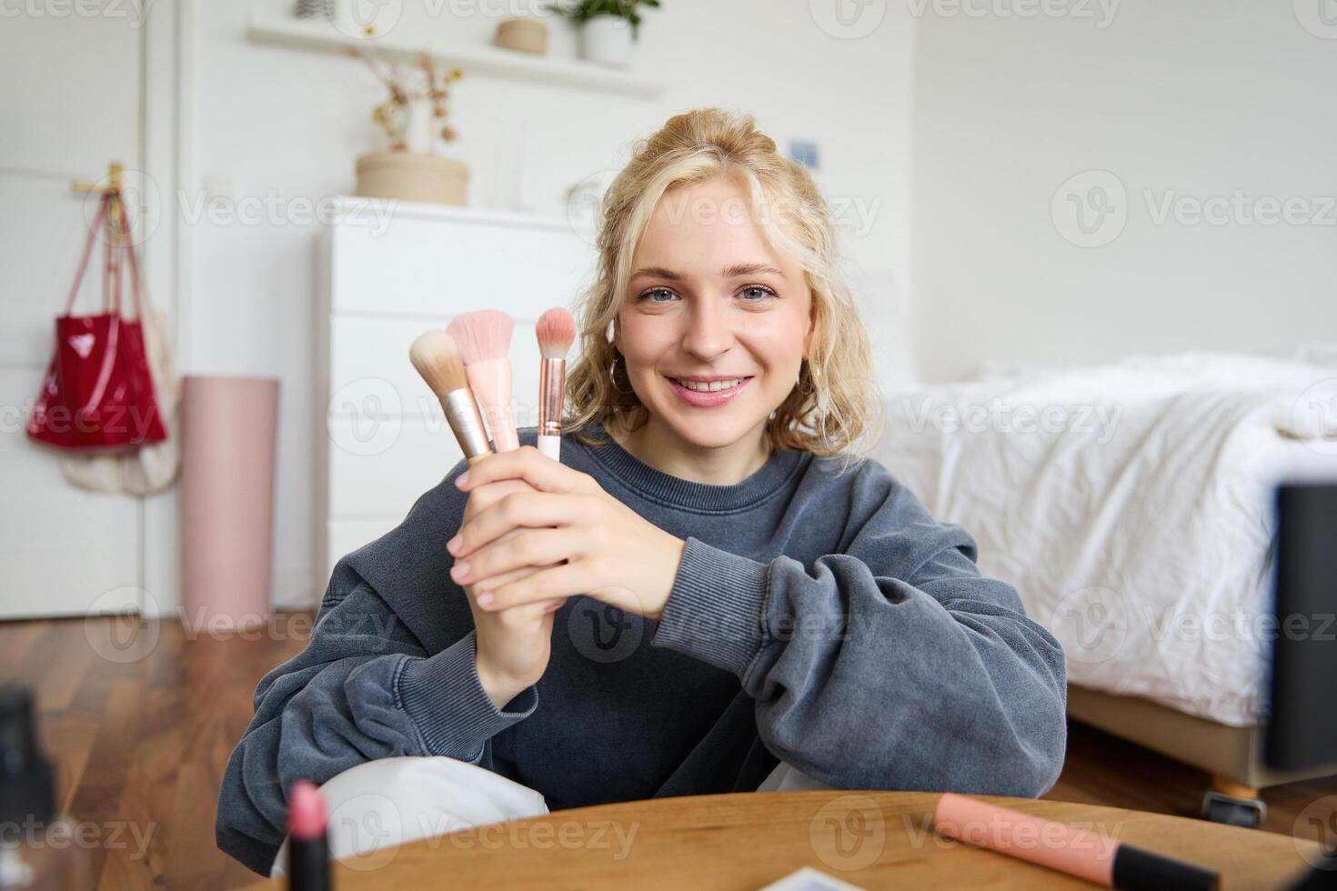 Portrait of young woman, content creator, making a about makeup, showing brushes to audience, looking at camera, recording beauty tutorial, smiling happily photo