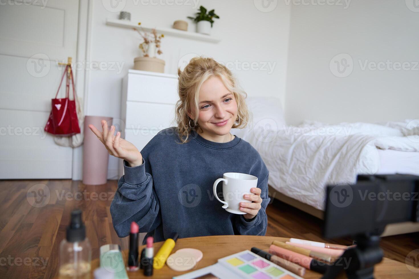 Image of young woman, makeup vlogger, sitting in bedroom with digital camera, drinking tea and talking, creating lifestyle , social media content photo