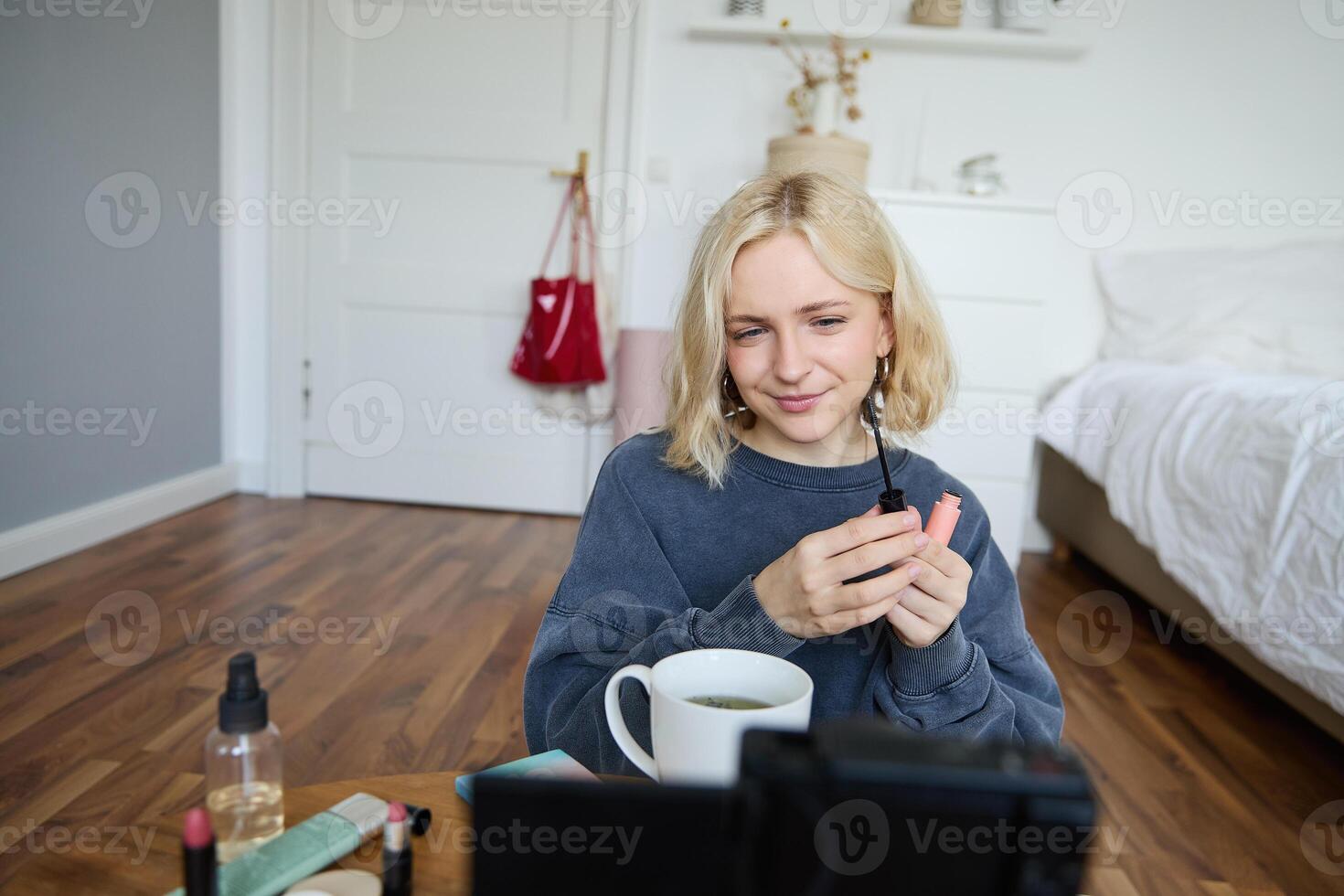 Close up portrait of young lifestyle blogger, woman sits in front of camera, looks thoughtful, applies makeup, holds mascara, explains how to put on beauty product for social media followers photo