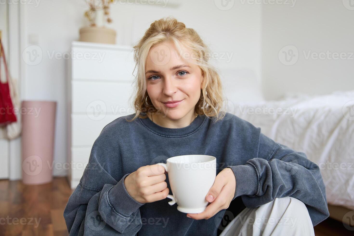 Portrait of young woman sitting on bedroom floor, drinking tea, holding white mug and smiling at camera photo