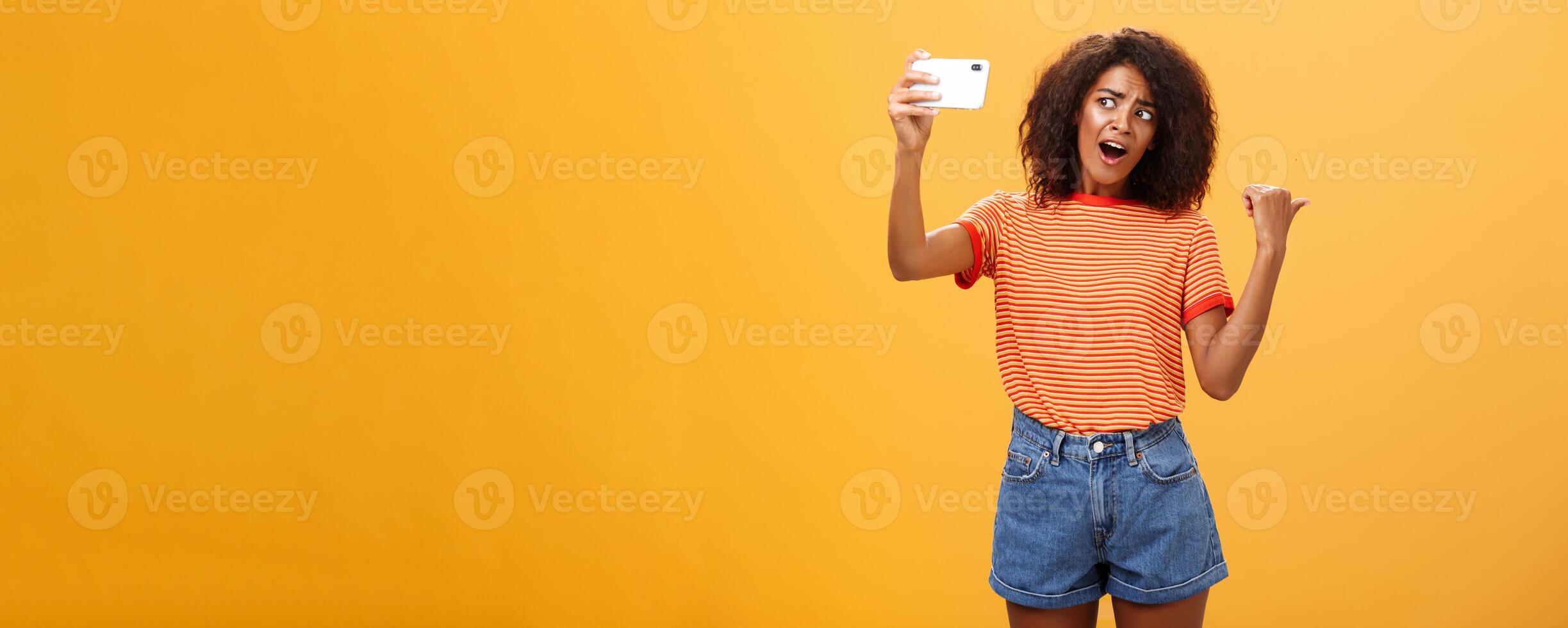 Woman recording blog pointing at strange object behind her. Portrait of concerned and curious stylish famous internet star holding smartphone talking in cellphone camera over orange background photo