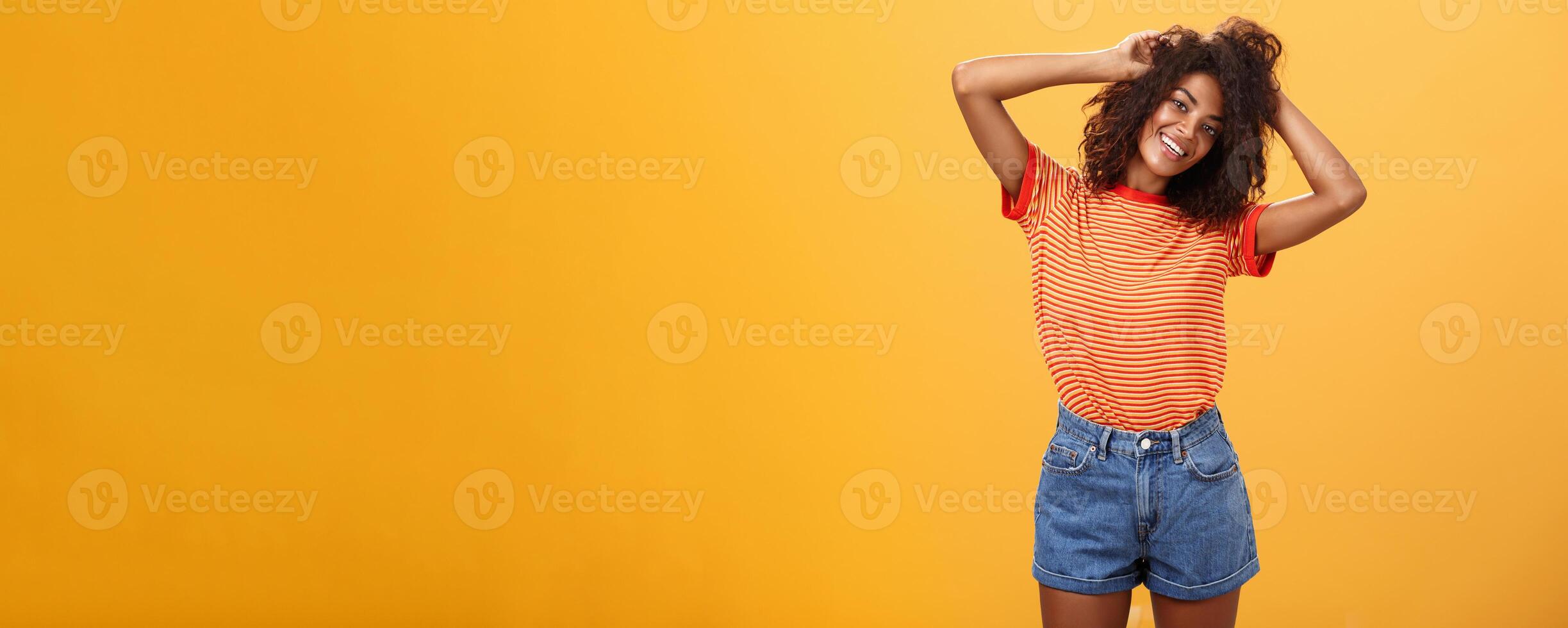 Time start living life fullest. Joyful optimistic woman having fun during vacation tilting head touching curly hair and enjoying summer sunshine in trendy striped t-shirt and shorts over orange wall photo