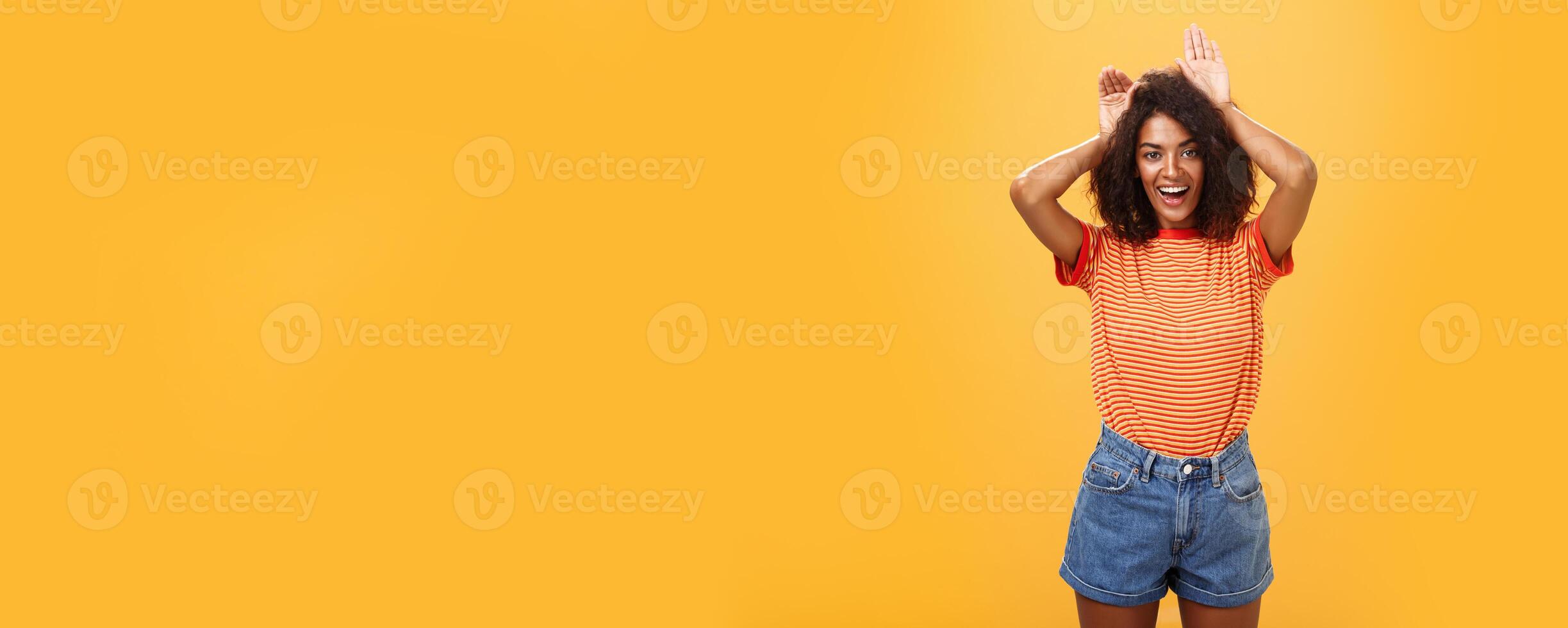 Let me be your bunny. Portrait of charming enthusiastic and charismatic happy dark-skinned female with afro hairstyle holding palms on head like animal ears making cute face over orange background photo